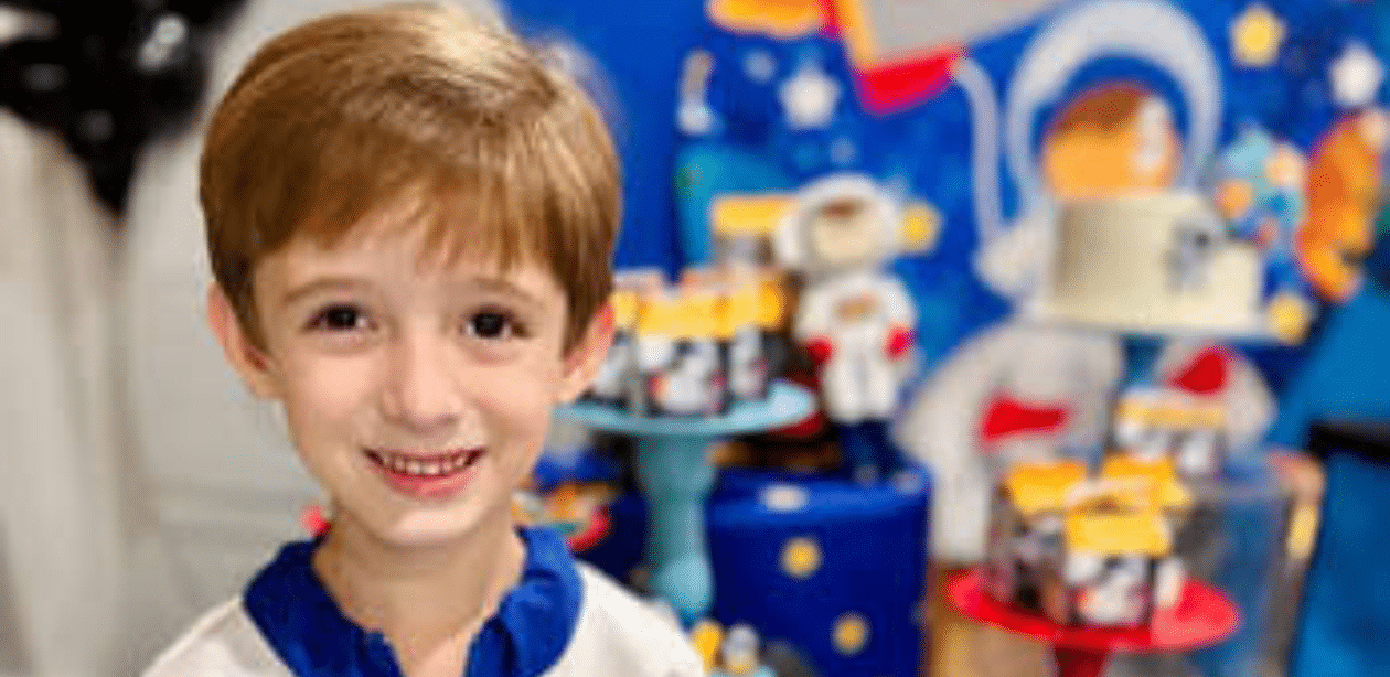 Menino superdotado de 5 anos sorri em festa temática, com decoração infantil ao fundo representando seu interesse por astronomia.