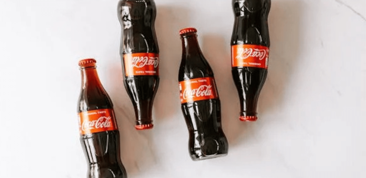 Cinco garrafas de vidro de Coca-Cola dispostas sobre uma superfície branca, algumas em pé e outras deitadas