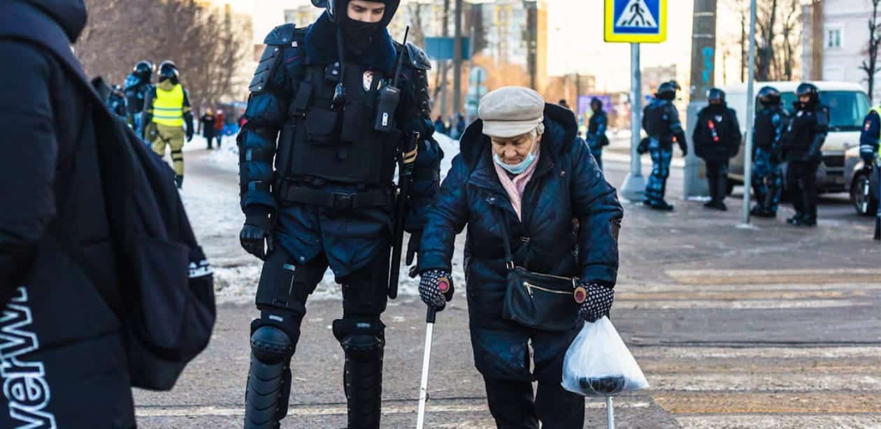 Policial equipado auxilia idosa a atravessar a rua em um cenário urbano. A idosa caminha com o auxílio de uma bengala e carrega uma sacola, enquanto o policial, vestindo uniforme tático, a acompanha. Ao fundo, outros agentes estão posicionados, indicando um ambiente de segurança e apoio à comunidade