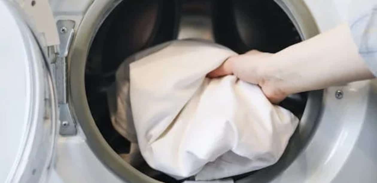 Mão colocando roupas brancas dentro de uma máquina de lavar roupa de abertura frontal.