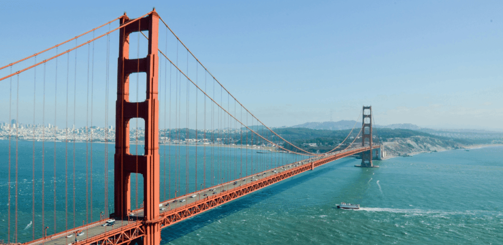 A imagem mostra a famosa Ponte Golden Gate, localizada na Califórnia, EUA. A ponte, com sua estrutura vermelha icônica, se estende sobre as águas do oceano, conectando a cidade ao condado de Marin. Ao fundo, é possível ver a linha do horizonte de San Francisco, com suas colinas e a costa ao longe. A cena é clara e ensolarada, com um barco navegando nas águas calmas, criando uma vista panorâmica impressionante e destacando a grandiosidade da ponte.