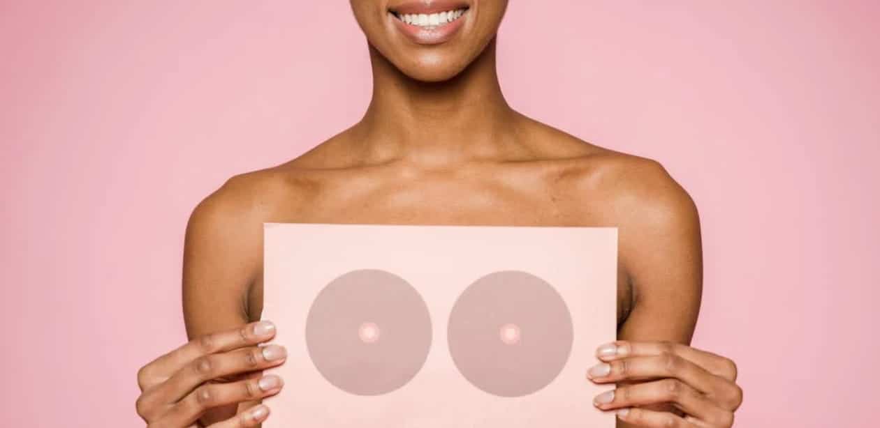 Mulher sorridente segura um cartaz com ilustração de seios sobre o peito, em fundo rosa, representando a autoestima feminina e a reconstrução após a mastectomia.