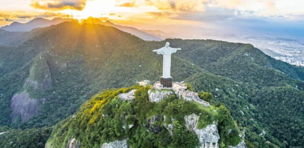 Vista aérea do Cristo Redentor, um dos principais cartões-postais do Brasil, no alto do Morro do Corcovado, cercado por uma exuberante paisagem verde. O pôr do sol ilumina a cena, destacando a beleza natural do Rio de Janeiro e a grandiosidade do monumento