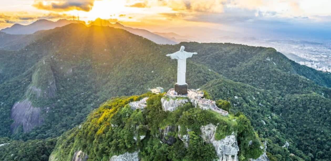 Vista aérea do Cristo Redentor, um dos principais cartões-postais do Brasil, no alto do Morro do Corcovado, cercado por uma exuberante paisagem verde. O pôr do sol ilumina a cena, destacando a beleza natural do Rio de Janeiro e a grandiosidade do monumento