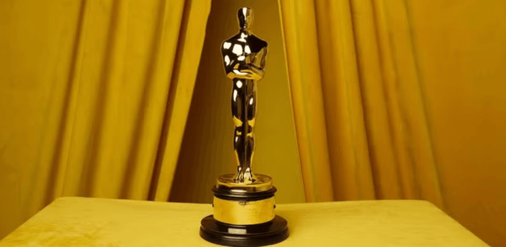 Estatueta dourada do Oscar em destaque sobre mesa amarela, com fundo de cortinas douradas.