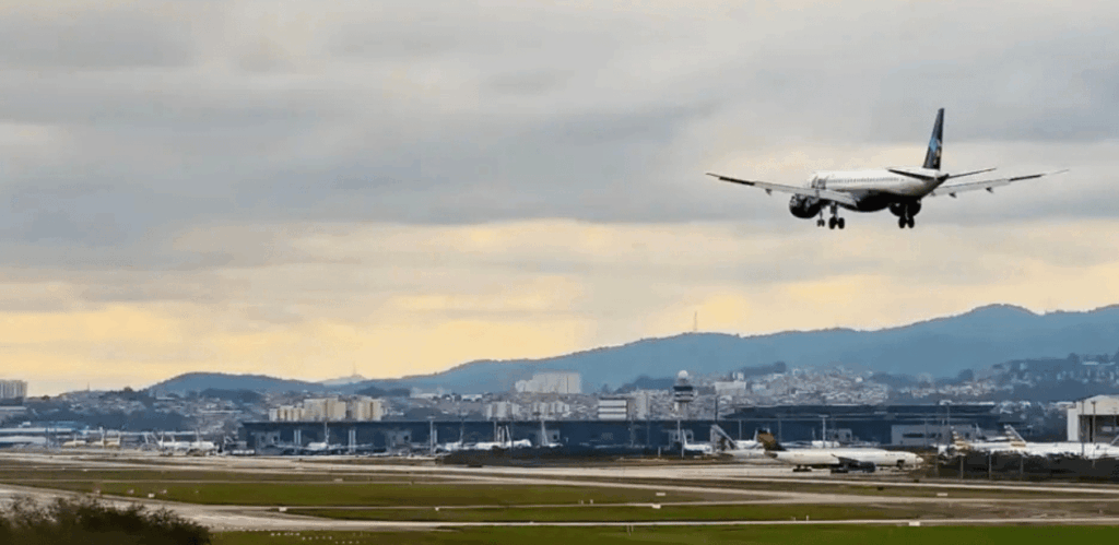 Avião se aproximando para pouso na pista do Aeroporto de Guarulhos, com terminal e cidade ao fundo