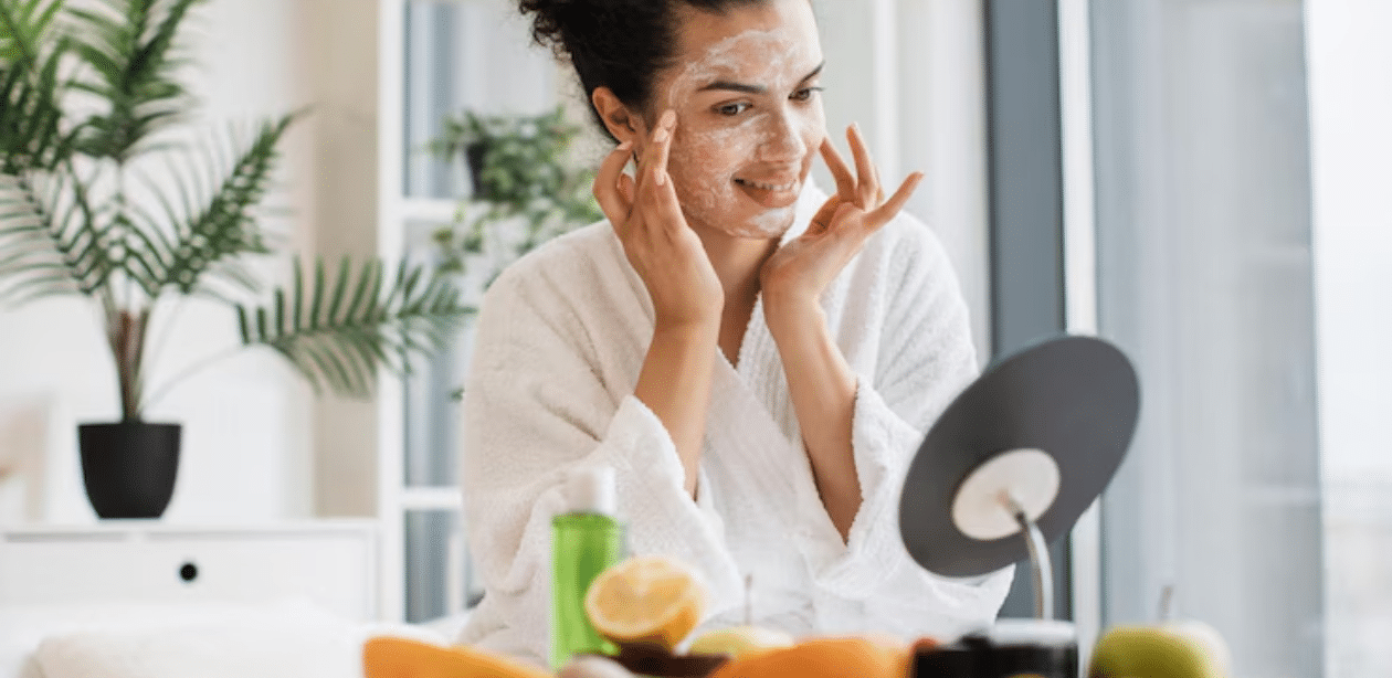 Mulher jovem aplicando máscara facial enquanto sorri para o espelho, com frutas frescas sobre a mesa
