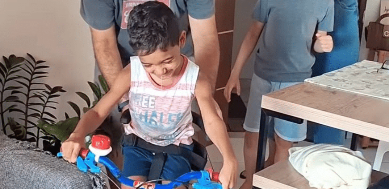 Menino com paralisia cerebral sorri ao receber bicicleta adaptada dentro de casa, em momento de inclusão e alegria.