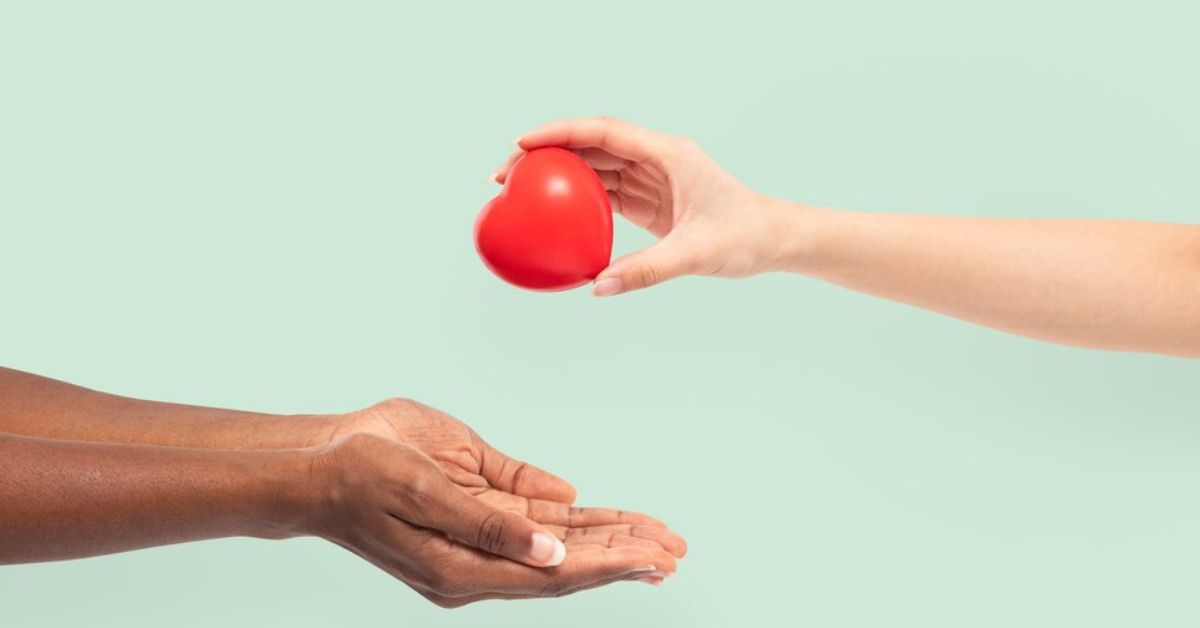 Mão branca entrega um coração vermelho simbólico para mãos negras estendidas, representando doação de órgãos com fundo verde claro.