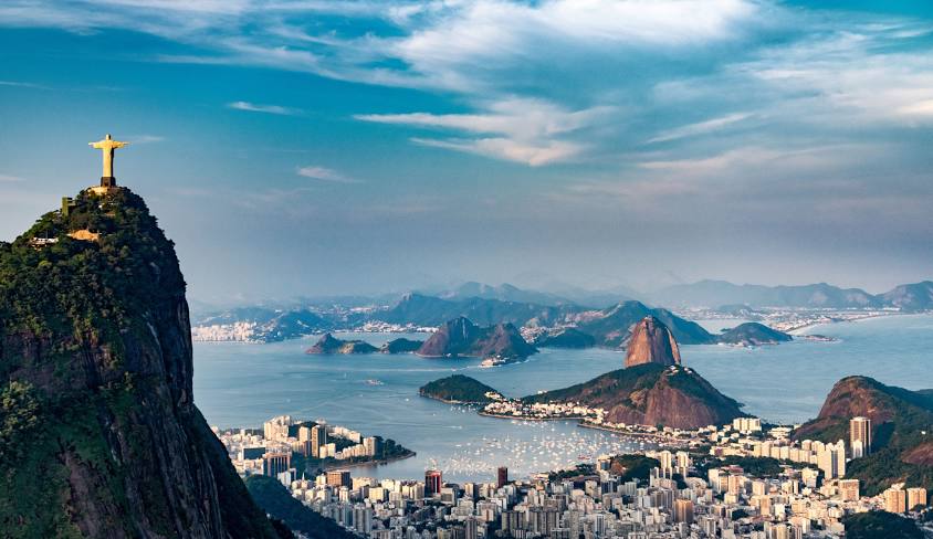 Vista panorâmica do Rio de Janeiro com o Pão de Açúcar e a Baía de Guanabara ao fundo, sob um céu parcialmente nublado.