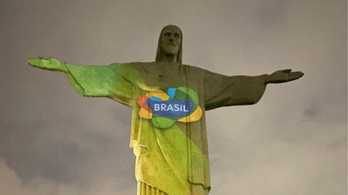 A imagem mostra o Cristo Redentor, um dos principais cartões-postais do Brasil, iluminado à noite com projeções coloridas da nova identidade visual da Embratur. No centro da estátua, aparece o logotipo com a palavra "Brasil", representando a promoção do turismo nacional em ações internacionais como o lançamento da nova plataforma Brasil Travel Specialist.