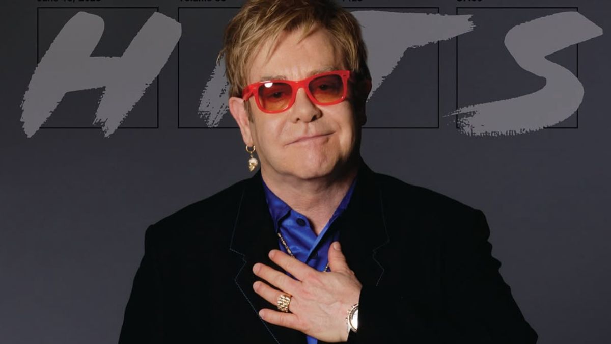 Elton John comemora sobriedade com gesto emocionado, vestindo óculos vermelhos e sorrindo diante de painel com a palavra “HITS”.