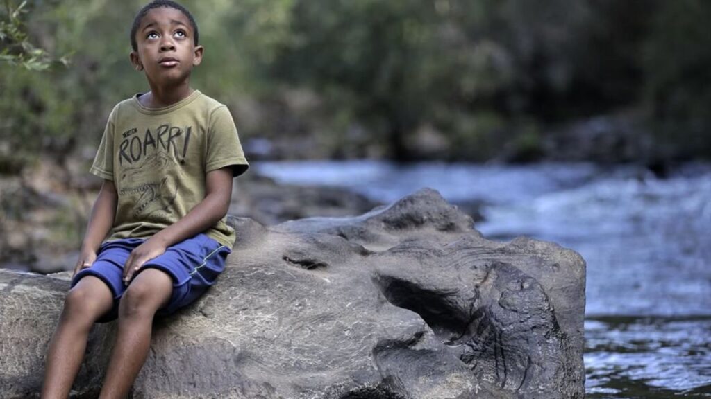 Menino quilombola sentado sobre uma pedra às margens do rio, em cena do filme sobre quilombolas “A Mensagem de Jequi”.