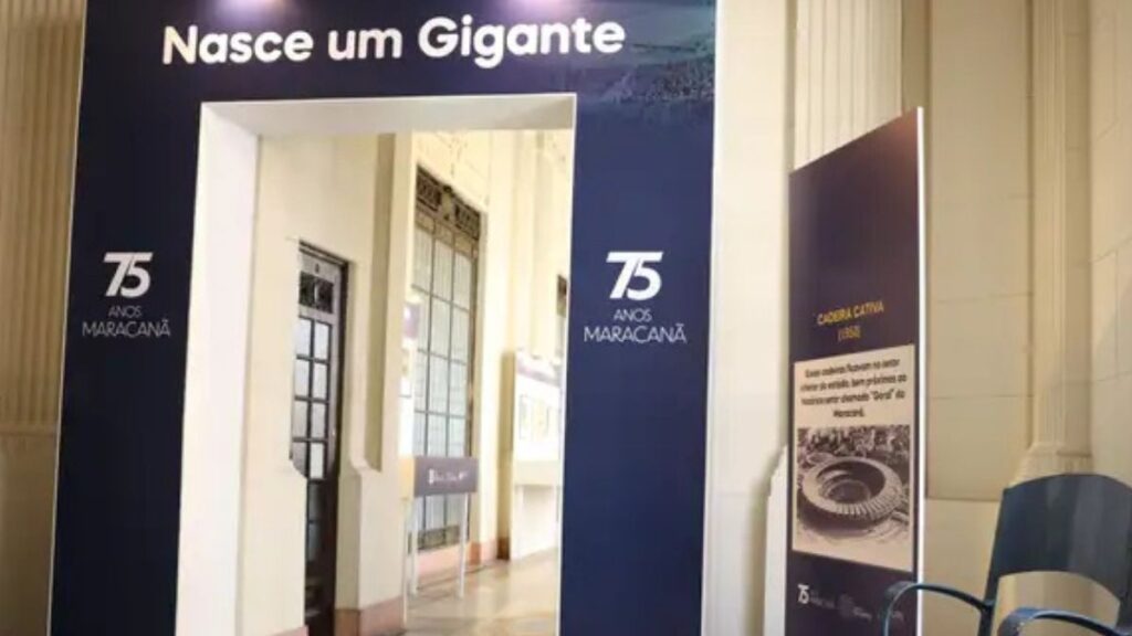 história do Maracanã Na foto vemos parte da exposição “75 Anos do Maracanã”, montada no Palácio Tiradentes, no Rio de Janeiro. O pórtico azul com a frase “Nasce um Gigante” recebe os visitantes, marcando a narrativa da inauguração do estádio em 1950. Ao lado, em destaque, está uma das peças simbólicas do acervo: uma cadeira cativa da época da inauguração, objeto histórico que remete ao privilégio de torcedores que tinham assento garantido no estádio mais famoso do Brasil.