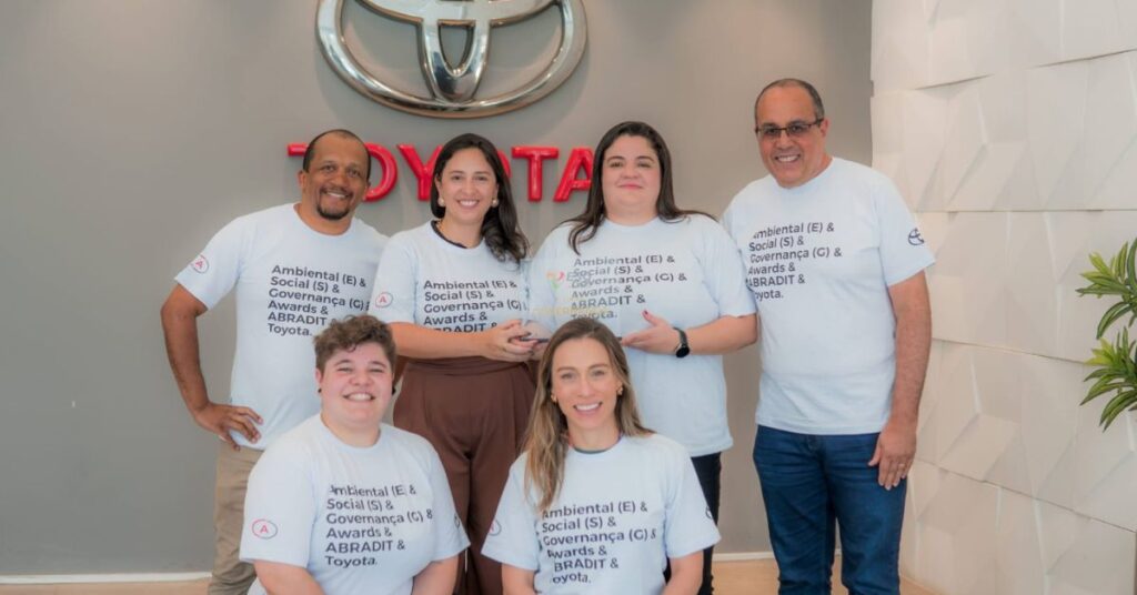 Colaboradores do Grupo New posam em frente ao logotipo da Toyota, usando camisetas temáticas de ESG, após premiação da ABRADIT & Toyota.