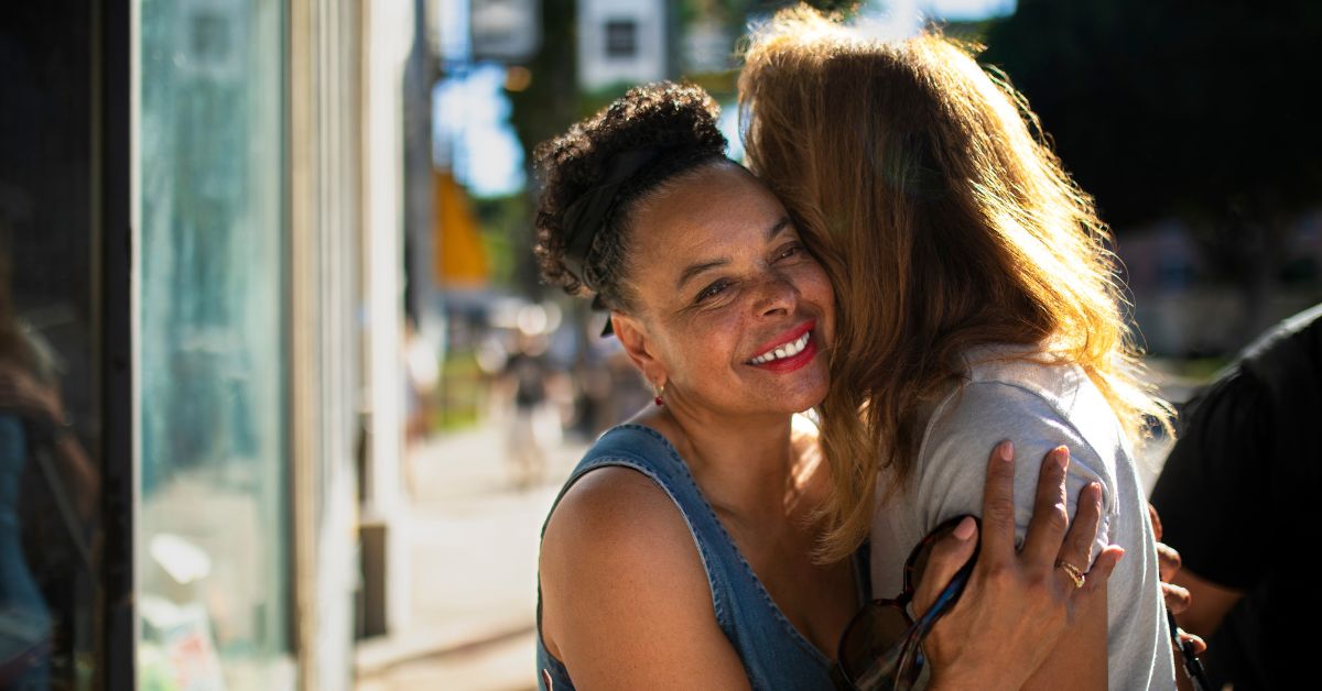 Convivência entre gerações reforça vínculos de afeto e respeito. Descrição: Duas pessoas de gerações diferentes se abraçam sorrindo em um ambiente urbano iluminado.