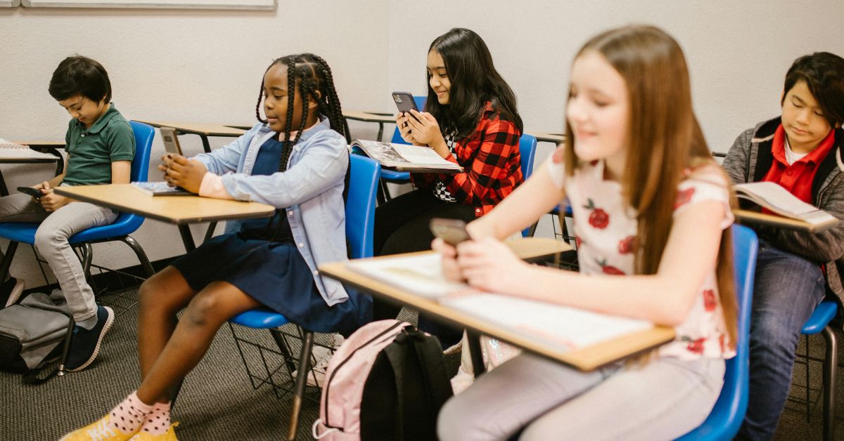 Crianças usando celular em sala de aula, ilustrando debates sobre segurança para crianças nas redes.