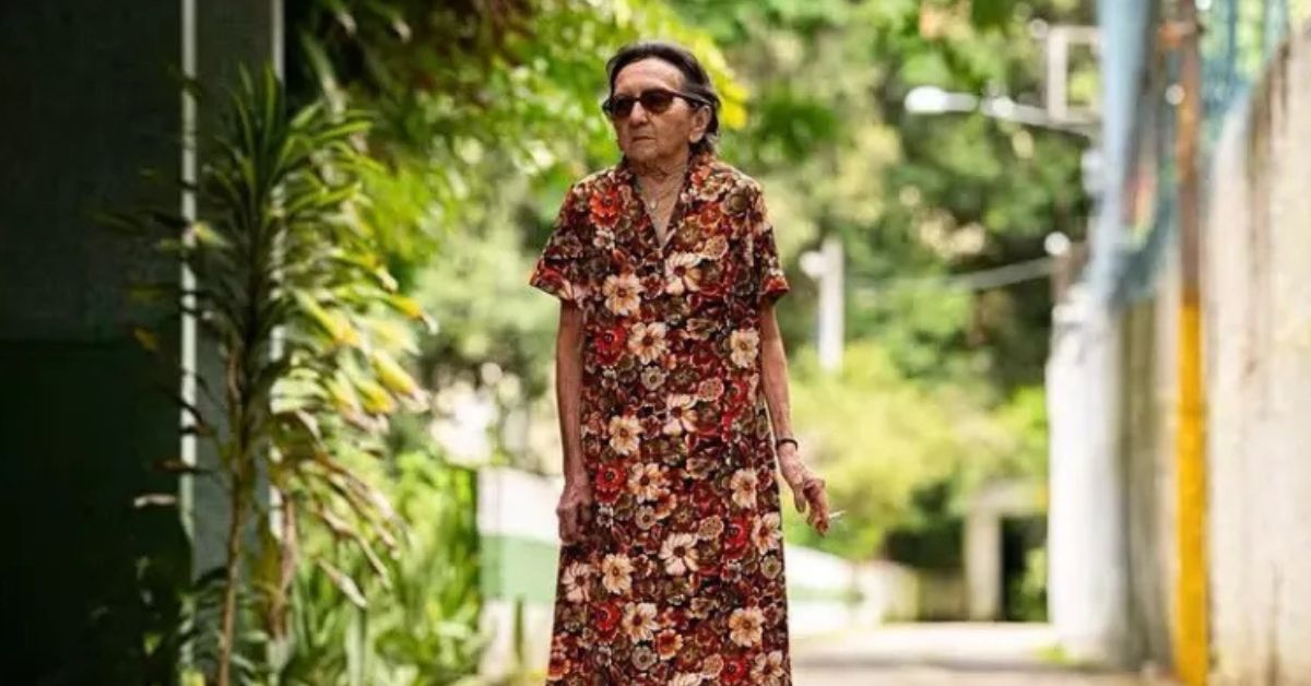 Tânia Maria caminha em cena ao ar livre em O Agente Secreto, usando vestido floral.