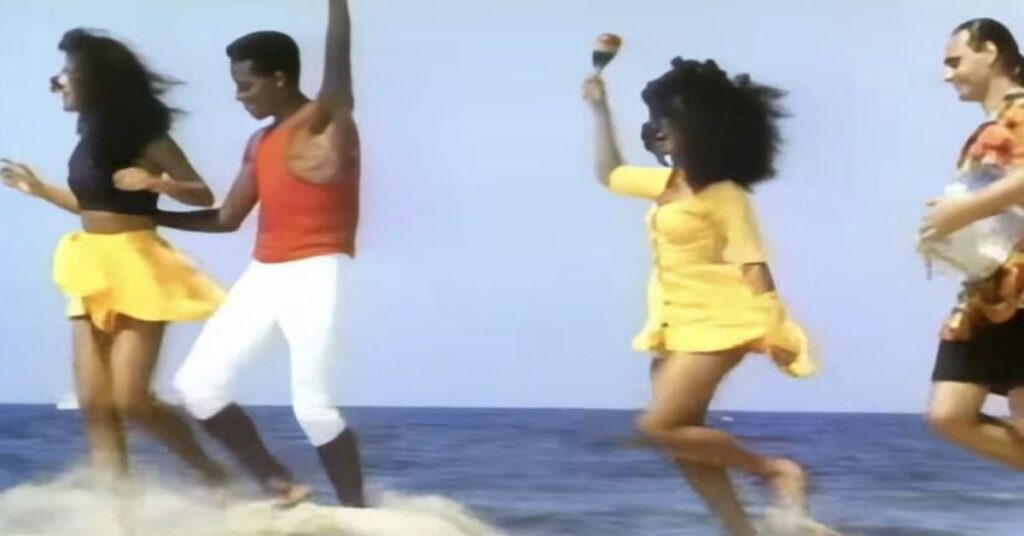Dançarinos apresentam o ritmo lambada em cenário de praia durante clipe dos anos 1980